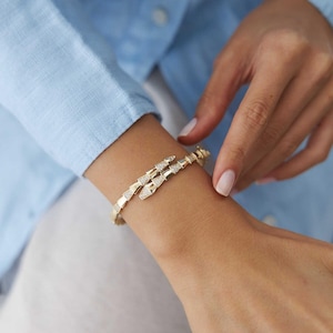 Könnte beinhalten: Ein goldenes Armband mit flexiblem, segmentiertem Design, das sich um das Handgelenk legt. Das Armband besteht aus mehreren dreieckigen Gliedern, wobei einige der Segmente mit kleinen, klaren, funkelnden Steinen besetzt sind.