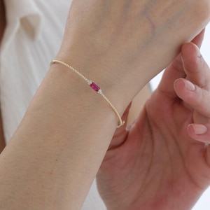Puede incluir: Una delicada pulsera de cadena dorada con una piedra preciosa de rubí rectangular de corte baguette en el centro, flanqueada por dos pequeñas piedras redondas transparentes a cada lado. La pulsera tiene un diseño de cadena trenzada.