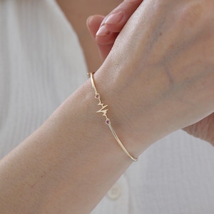 Puede incluir: Una pulsera rígida de oro fino con un diseño central de línea de electrocardiograma. El motivo del latido del corazón está flanqueado por dos pequeñas piedras preciosas redondas de color rosa engastadas en la banda de oro pulido y brillante.