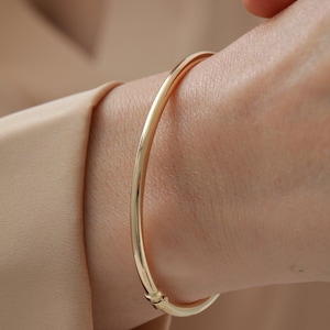 May include: A polished gold bangle bracelet with a smooth, rounded surface and a visible hinged clasp mechanism. The bracelet features a continuous, circular design with a reflective metallic finish, worn around the wrist area.