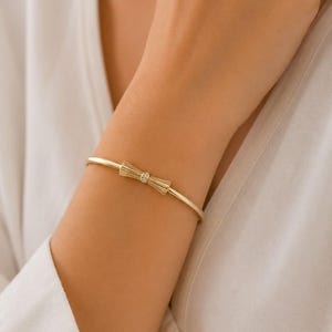 Pode incluir: Uma pulseira rígida fina em ouro com um ornamento central em forma de laço. O laço tem uma textura estriada e é decorado com uma pequena pedra clara e brilhante no centro. A pulseira tem um acabamento liso e polido com forma circular.