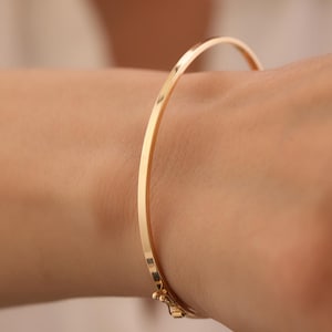 Puede incluir: Una pulsera tipo brazalete fina de oro pulido con una superficie lisa y redondeada y un mecanismo de cierre de bisagra seguro. El metal tiene un tono amarillo cálido y reflectante, creando una forma circular simple y continua.
