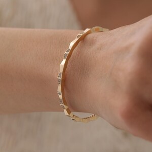 Może przedstawiać: Złota bransoletka typu bangle o geometrycznym, fasetowanym wzorze z małymi, osadzonymi przezroczystymi kamieniami wzdłuż zewnętrznych krawędzi każdego segmentu. Bransoletka ma polerowane metaliczne wykończenie i ciągłą, okrągłą strukturę.