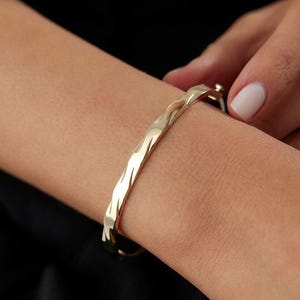Könnte beinhalten: Ein goldfarbener Armreif mit einer polierten, reflektierenden Oberfläche und einer ausgeprägten wellenförmigen Textur entlang der gesamten Länge. Der Armreif verfügt über einen seitlich sichtbaren Klappverschluss, der eine geschlossene Form bildet.