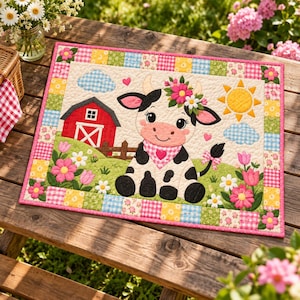 Puede incluir: Un tapete de tela acolchada rectangular con una ilustración central de una vaca blanca y negra que lleva una corona de flores y un pañuelo rosa con estampado de corazones. La escena incluye un granero rojo, un sol amarillo y flores.