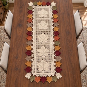 May include: A rectangular crochet table runner featuring a central grid pattern with five repeating cream-coloured maple leaf motifs. The border is decorated with a continuous row of small, multi-coloured maple leaves in shades of red, orange, yellow, brown, and green.