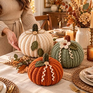 May include: Three crocheted pumpkins in cream, sage green, and burnt orange. Each pumpkin features a dark green stem, small wooden buttons, and decorative crocheted leaves or vines. The pumpkins are textured with vertical ribbed stitching.