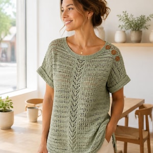 May include: A sage green short-sleeved knit top featuring an openwork stitch pattern, a vertical chevron design down the center, and three round brown buttons on the left shoulder. The garment has a round neckline and cuffed sleeves.