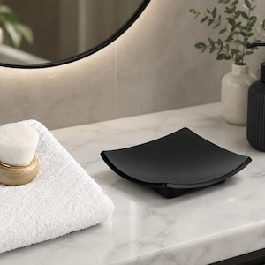 May include: A square, matte black soap dish with curved, raised edges. The dish has a smooth, concave surface designed to hold a bar of soap. It is positioned on a white marble surface next to a white textured towel and a wooden facial brush.