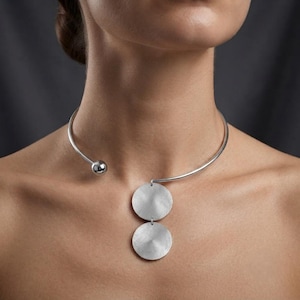 May include: A silver-toned metal choker necklace featuring a smooth, open-ended band with a small spherical terminal on one side. A vertical pendant hangs from the center, consisting of two brushed, circular metal discs linked together in a row.