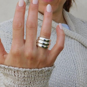 Può includere: Un anello in argento lucido caratterizzato da un design a tripla fascia con tre strati impilati, lisci e arrotondati. L'anello presenta una finitura metallica molto brillante e una forma continua e uniforme che avvolge il dito, creando un aspetto stratificato.
