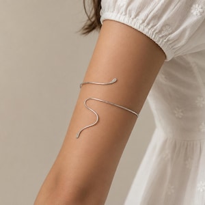 May include: A silver-toned metal arm cuff featuring a slender, continuous wire design that wraps around the upper arm in a fluid, serpentine shape with smooth, rounded ends. The piece has a polished, reflective finish and an open, adjustable structure.