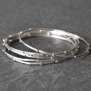 Może przedstawiać: Zestaw trzech cienkich, okrągłych srebrnych bransoletek typu bangle o matowym wykończeniu. Każda bransoletka posiada małe, równomiernie rozmieszczone wypukłe srebrne kropki wzdłuż zewnętrznego obwodu, co nadaje metalowym obręczom teksturę.