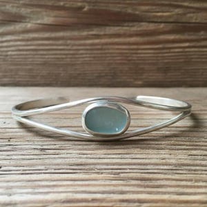 Puede incluir: Una pulsera tipo brazalete de tono plateado con una banda de metal lisa y pulida que se divide en dos líneas paralelas. En el centro, una piedra única de forma ovalada y color azul pálido está engastada en un marco de metal sencillo.