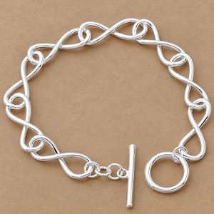 Op de afbeelding: Een zilverkleurige armband bestaande uit meerdere in elkaar grijpende, oneindig gevormde schakels. Het sieraad is voorzien van een knevelsluiting bestaande uit een cirkelvormige ring en een rechte staaf, wat zorgt voor een veilige sluiting van deze gepolijste metalen ketting.
