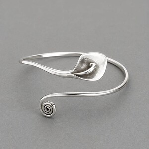Puede incluir: Un brazalete abierto de tono plateado con acabado pulido. Un extremo del brazalete tiene forma de flor de cala, mientras que el extremo opuesto termina en un pequeño diseño de espiral apretada que se curva hacia el centro de la pieza.