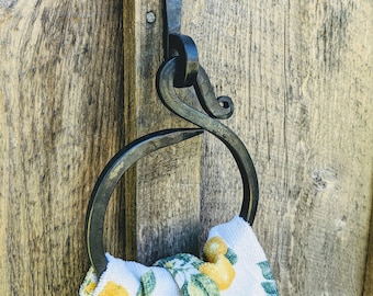 May include: A black metal towel ring with a decorative swirl design is mounted on a wooden wall. A white kitchen towel with a lemon print hangs on the ring.
