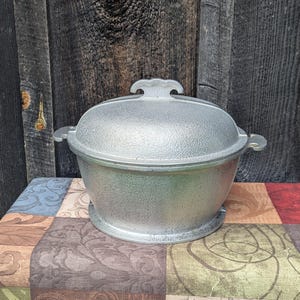 May include: A silver-colored metal pot with a textured finish and a matching lid. The pot features two side handles and a central handle on the lid. The exterior surface of the pot and lid has a consistent, slightly rough, granular appearance.