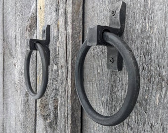 May include: Two black metal door handles with a ring design. The handles are mounted on a wooden door.