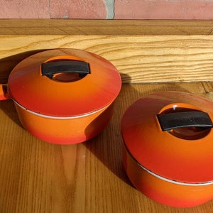 May include: Two orange enameled cast iron saucepans with lids. Each pan has a long handle and a black handle on the lid. The pans are on a wooden surface, with a brick wall in the background. The pans are a vibrant orange color.
