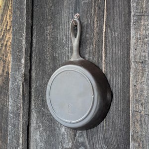 May include: A cast iron skillet with a handle, hanging on a weathered wooden surface. The skillet is dark gray and has a circular base with the number "3" and other markings. The handle has a hole for hanging.