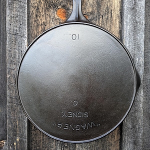 May include: A black cast iron skillet with a handle, resting on a wooden surface. The skillet is round with the text "WAGNER" and "SIDNEY O." and the number "10" embossed on the bottom. The pan is approximately 10 inches in diameter.