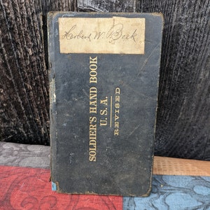 Puede incluir: Un libro encuadernado en cuero negro titulado "SOLDIERS HAND BOOK U.S.A. REVISED" con una etiqueta manuscrita en la portada que dice "Herbert W. Beck"