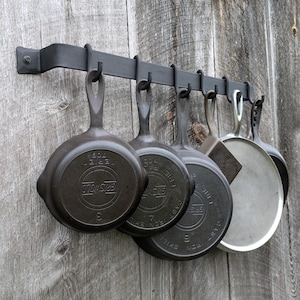 May include: A black metal pot rack with several cast iron skillets and a spatula hanging from it. The skillets are various sizes and have the brand name "Griswold" on them. The rack is mounted on a weathered wooden wall.