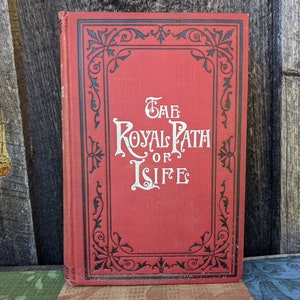 以下が含まれることがあります： 黒と白の装飾的な枠で飾られた赤いハードカバーの本。本のタイトルは「The Royal Path of Life」です。
