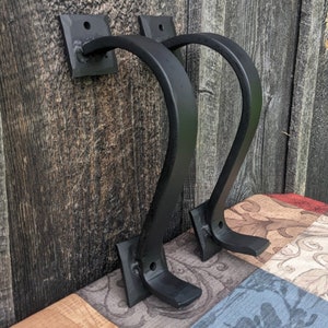 Puede incluir: Dos ganchos de pared de metal negro con un diseño curvo. Los ganchos están montados en una pared de madera y están listos para colgar objetos.