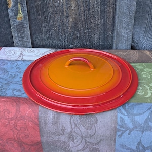 Pode incluir: Tampa de panela redonda de ferro fundido esmaltado, com um centro laranja brilhante que se desvanece para uma borda vermelha vibrante. A tampa tem uma alça curva e é colocada sobre um tecido estampado e um fundo de madeira envelhecida.