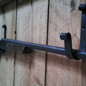 May include: A dark metal rifle-shaped wall mount, mounted on a wooden surface. The mount has a long, cylindrical barrel and several curved brackets for holding a rifle. The wood has vertical lines.