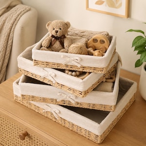 May include: A set of three rectangular woven wicker storage baskets stacked on top of each other. Each basket features a light beige fabric liner with a small fabric bow attached to the front center. The top basket contains a teddy bear.