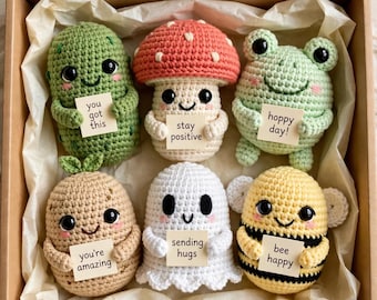 Pode incluir: Um conjunto de seis figuras de crochê em uma caixa de papelão, incluindo um picles verde, um cogumelo vermelho, um sapo verde-claro, uma batata bege, um fantasma branco e uma abelha amarela, cada um segurando uma pequena placa com mensagens.