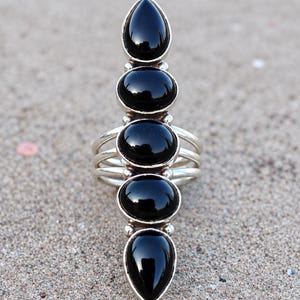 May include: A silver ring featuring five vertically aligned, smooth black stones. The design includes two teardrop-shaped stones at the top and bottom, with three oval-shaped stones in the center, all set in polished silver with a multi-band shank.
