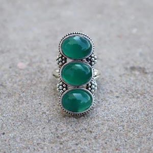 May include: A silver ring featuring three vertically aligned oval green cabochon stones. The metal setting has a decorative beaded texture surrounding each stone and along the sides of the band, creating a detailed and ornate appearance.