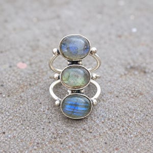 May include: A silver ring featuring three vertically stacked oval labradorite gemstones. Each stone displays iridescent blue and green flashes set within a polished silver frame. The band consists of three separate silver wires that connect to the settings.