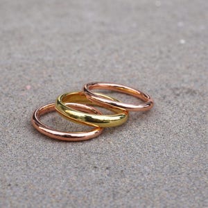May include: A set of three smooth, polished metal rings featuring a simple, rounded band design. The collection includes one ring in a bright yellow gold tone, one in a warm rose gold tone, and one in a copper tone, all stacked together.