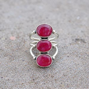 Puede incluir: Un anillo de plata con tres piedras preciosas de rubí rojo de forma ovalada, facetadas y alineadas verticalmente. La banda está formada por varios hilos de plata finos que se conectan a los engastes de cada piedra, creando un diseño abierto.