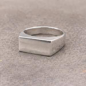 May include: A polished silver-toned ring featuring a wide, flat, rectangular face. The band is smooth and solid, with a clean, unadorned surface that wraps around the finger. The metal has a reflective finish, highlighting its simple, geometric shape.
