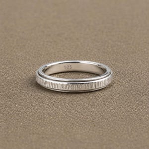Könnte beinhalten: Ein silberfarbener Metallring mit einem strukturierten Muster aus vertikalen Linien auf dem äußeren Band. Die Innenseite des Rings ist glatt und deutlich mit dem Stempel 925 versehen, was auf eine Zusammensetzung aus Sterlingsilber für dieses Schmuckstück hindeutet.