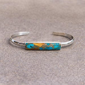 May include: A silver cuff bracelet featuring a rectangular inlay of turquoise stone with vibrant blue and golden-brown matrix patterns. The metal band is polished and smooth, curving into an open design that allows for easy placement on the wrist.