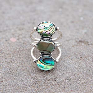 May include: A silver ring featuring three vertically stacked oval abalone shell cabochons. The top stone displays iridescent green and pink hues, the middle stone is a muted olive green, and the bottom stone shows vibrant blue and green tones.