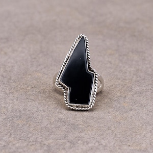 Op de afbeelding: Een zilveren ring met een asymmetrische, gepolijste zwarte steen. De steen is omlijst door een gedraaide, touwachtige zilveren rand, wat zorgt voor een opvallende geometrische vorm. De ringband is gemaakt van glad, gepolijst zilver.