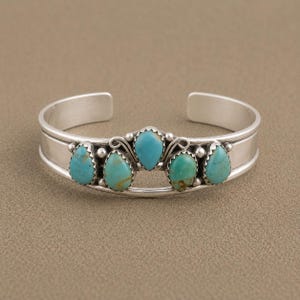 May include: A silver cuff bracelet featuring five teardrop-shaped turquoise stones arranged in a row. The stones are set in serrated bezels with small silver bead accents between them. The band is smooth, polished silver with an open back design.