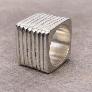 May include: A square-shaped silver ring featuring a series of parallel, raised vertical ridges across its outer surface. The band has a polished metallic finish with a hollow center, showcasing a distinct geometric design with clean, sharp edges.