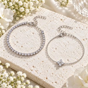 Peut inclure: Deux bracelets en métal argenté ornés de pierres transparentes taille ronde. L'un présente une ligne continue de pierres, tandis que l'autre possède une chaîne fine avec un pendentif en forme de fleur à quatre pétales en pierres.