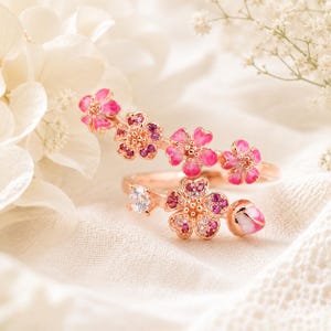 May include: A rose gold bypass ring featuring multiple cherry blossom motifs. The floral elements are accented with pink and red gemstones, white enamel detailing, and a single clear round stone on one end, creating a delicate, layered appearance.