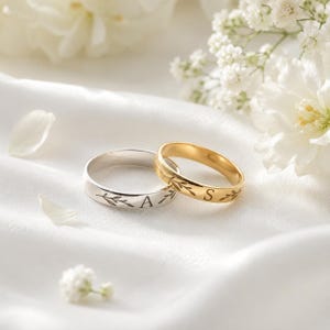 Può includere: Due anelli in metallo, uno color argento e uno color oro, ciascuno caratterizzato da un motivo a foglie inciso e da una singola lettera. L'anello argento è inciso con la lettera A, mentre l'anello oro è inciso con la lettera S. Entrambi sono lisci.
