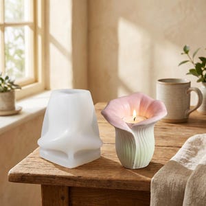 May include: A translucent white silicone mold with a geometric shape sits next to a finished flower-shaped candle holder. The holder features a pale green base that transitions into soft pink petals, containing a small lit tea light candle.